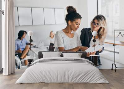 Slim black girl in jeans explaining something to european female colleauge while asian man talking with fair-haired young lady. Portrait of managers of international company solving work problems. Wall mural