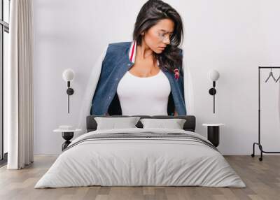 Slim dark-haired girl in white tank-top looking down holding hands in pocket. Indoor portrait of brunette female model in glasses and trendy bomber jacket. Wall mural