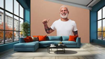 Smiling adult man looks into camera on beige background. Handsome guy with gray beard in white T-shirt is smiling and showing thumb up Wall mural