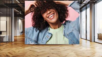 Smiling curly woman in denim jacket posing over pink background. Joyful happy girl in green tee laughing on isolated backdrop Wall mural