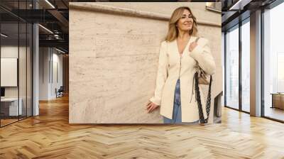 Smiling cute caucasian adult lady in beige coat and jeans posing against wall on street. Blonde with long hair walks alone in city. Lifestyle, concept Wall mural