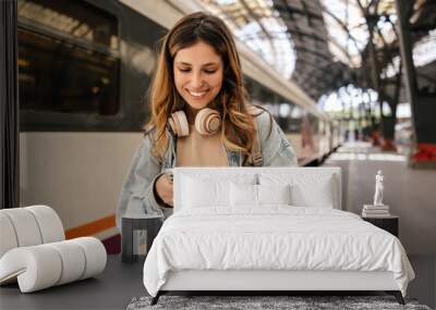 Smiling young caucasian lady is registering on site for buying tickets standing at station. Blonde woman with wavy hair wears casual clothes. Concept of use Wall mural