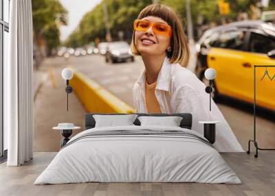 Smiling young caucasian woman looking at camera walking in public place. Brown-haired with bob haircut wears white shirt, orange glasses. Good mood concept Wall mural