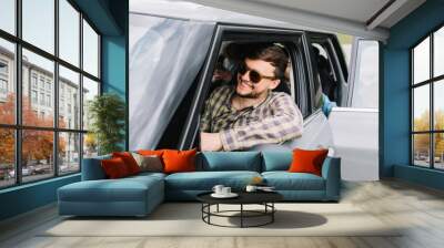 Smiling young man with trendy haircut and beard traveling with family around Italy by car. Laughing driver in stylish dark sunglasses ready to continue journey with friends in vacation. Wall mural