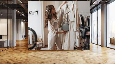 Stilysh woman in white outfit holding beige stylish trench coat. Curly pretty lady in fashionable pants and blouse poses with handbag in apartment Wall mural