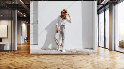 Stylish girl in gray pants and white cotton blouse posing near white wall. Photo of woman in cap and glasses Wall mural