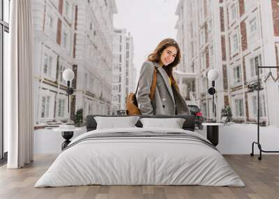 Stylish lady with brown backpack walking around city under snowfall. Outdoor photo of pretty girl with charming smile posing in gray coat on urban background in winter day. Wall mural