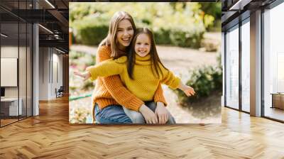 Stylish woman with dark hair holding daughter on her knees. Outdoor photo of glad young lady having fun in autumn park with child. Wall mural