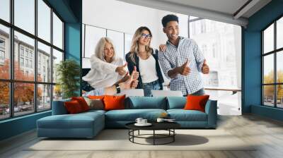 Tall african manager with big smile holding thumbs up and looking away. Charming girls working as managers posing in office next to table with laptops on it and laughing. Wall mural