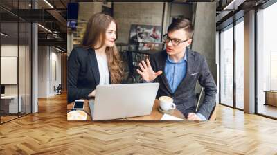 Two colleagues discuss something near laptop. Man in blue shirt and grey jacket describes something with hand gestures. Woman with long hair wearing black jacket and white blouse listens and smiles. Wall mural