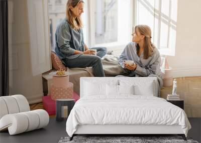 Two cute caucasian mother with her daughter drink delicious coffee at weekend sitting by window. Blondes in shirts and jeans spend their leisure time. Mood, lifestyle, concept. Wall mural