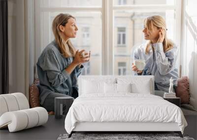 Two young caucasian girls friends enjoy conversation and drink coffee sitting on windowsill during day. Blondes wear casual denim. Positive human emotions concept Wall mural