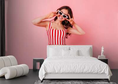 tylish dark-haired woman in summer shorts and top smiles and shows peace sign on pink background Wall mural