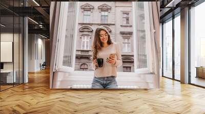 Winsome girl in casual attire spending time at home and posing on city background with cup of tea. Graceful lady in glasses texting message near window. Wall mural