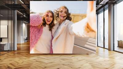 Winsome woman in pink coat having fun with friend. Blithesome lady in white attire making selfie with sister. Wall mural