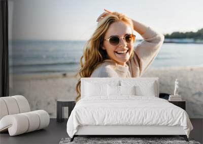 Winsome woman in sunglasses spending cold morning beside sea. Blithesome blonde lady with cup of coffee laughing at ocean coast Wall mural