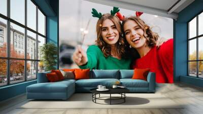 Women celebrating winter holidays. Outdoor photo of two girls with sparklers laughing at camera. Wall mural