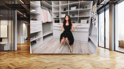 Young brunette woman sitting in a huge dressing room thinks over the choice of clothes, she is dressed stylish black outfit and silver shoes, expressing true positive face emotions. Wall mural