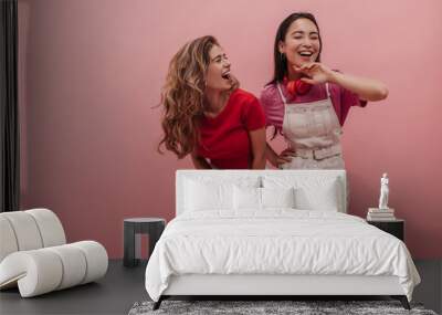 Young caucasian lady laughs hard with her pretty asian girlfriend against pink background. Image of 20s girls pretending dancing to music and having fun with each other in studio. Wall mural