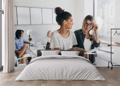 Young female executive explaines new strategy to blonde employee in glasses and smiling. Indoor portrait of multicultural collective working on project in office and using laptop. Wall mural