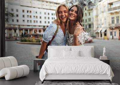 Young female friends in stylish jeans and floral trendy blouses hug, smile and pose in great mood at city square. Wall mural