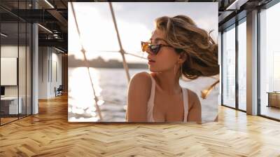 Young lady with fluffy blonde hair in modern beach outfit and dark sunglasses posing in view of sea at sunshine and looking away Wall mural