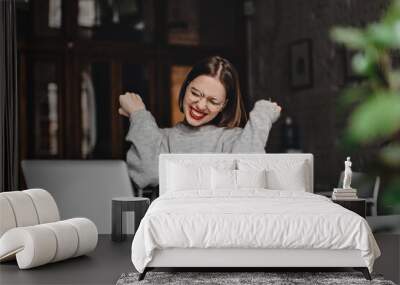 Young woman with red lipstick happily clenches fists and laughs. Portrait of lady wearing glasses with red lipstick working in laptop in cafe Wall mural