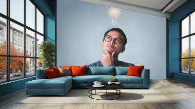 Thinking asian man in glasses looking up with light idea bulb above head on gray background.creative idea.Concept of idea and innovation Wall mural