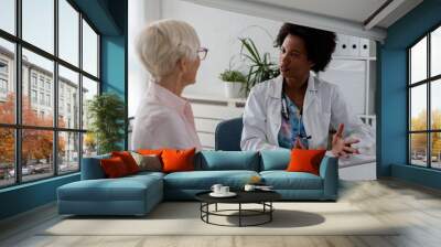 A female doctor sits at her desk and chats to an elderly female patient while looking at her  test results Wall mural