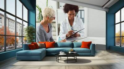 A female doctor sits at her desk and chats to an elderly female patient while looking at her  test results Wall mural