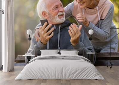 Active modern cheerful senior couple meet in the park. An older man is sitting on a park bench using smartphone, and an elderly woman surprises him from the back. Wall mural