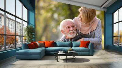 Active modern cheerful senior couple meet in the park. An older man is sitting on a park bench using smartphone, and an elderly woman surprises him from the back. Wall mural