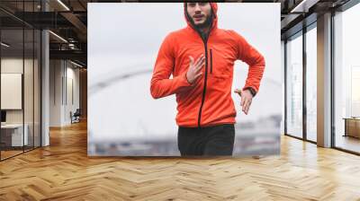 Handsome young athlete man running fast along river in orange windbreaker jacket. Extreame weather sport. Running on a rainy day. Wall mural