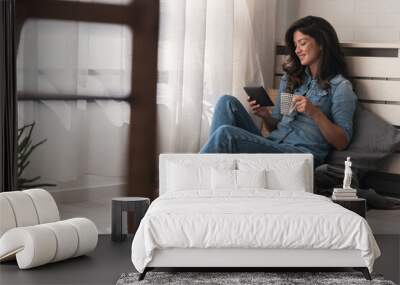 Handsome young woman reading an e-book, drinking coffee at home sitting cozy by the window Wall mural