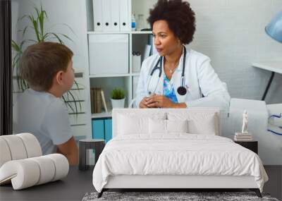 Smiling female afro american doctor general practitioner talks and amuses child before medical examination Wall mural