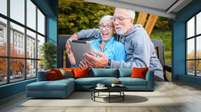 Smiling senior active couple sitting on the bench in the park looking at tablet computer. Using modern technology by elderly. Wall mural