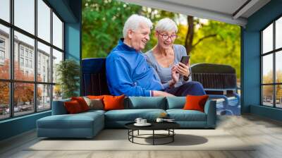 Smiling senior active couple sitting on the bench looking at smartphone Wall mural