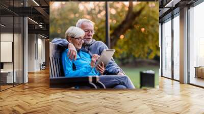 Smiling senior active couple sitting on the bench looking at tablet computer. Using modern technology by elderly. Wall mural