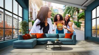 Three young women of diverse backgrounds joyfully holding hands and running down a sunny, palm-lined street in a vibrant, urban neighborhood. Their laughter and energy capture the spirit of friendship Wall mural