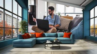 Young bearded businessman working on laptop computer while sitting on sofa at his home office Wall mural