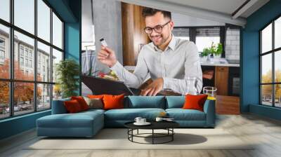 Young man holding credit card sitting in front of laptop computer at home, paying for online order. People, lifestyle, modern technologies and e-commerce concept. Online banking and shopping using mod Wall mural