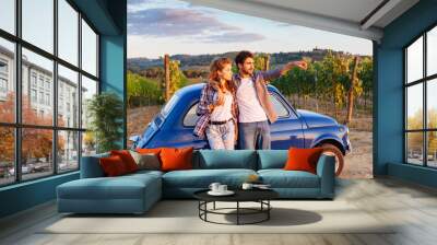 A loving couple watch the sunset, standing leaning against an old blue car in Tuscany, Italy. Around them, rows of vines and the classic hills. The young man shows to his girlfriend sunset Wall mural