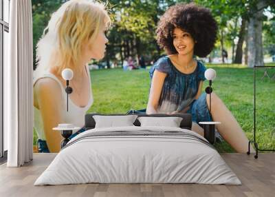 Couple of young women talk to each other sitting on the grass of a meadow on a summer day at the park - Millennials have fun together laughing and joking Wall mural