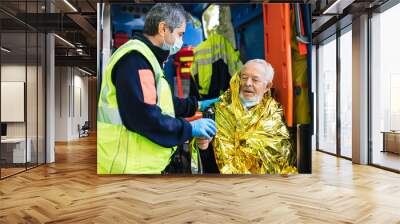 Paramedic rescues an elderly person in distress with an ambulance by covering him with a thermal blanket and making him drink a hot drink - Concept of first aid Wall mural