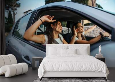 Two young women sing a song on the radio and dance in the car on a day trip in the summer - Best friends having fun together driving around the countryside - Smiling millennial in a relaxing moment Wall mural