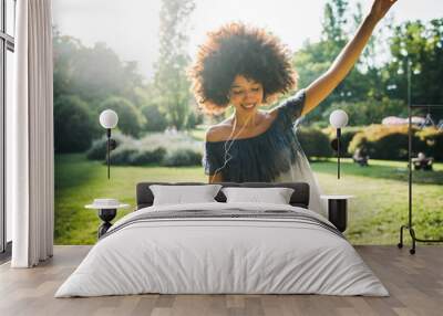 Young black woman dancing listening to the music with earphones from the smart phone in a park at sunset on a summer day Wall mural