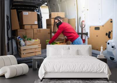 Young female courier unloads van for customer delivery of a basket of fruit and vegetables bought online at supermarket wearing face mask and gloves during global Coronavirus Covid-19 pandemic Wall mural