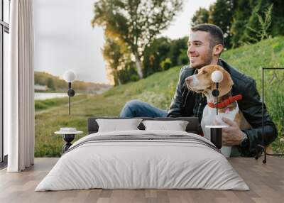 Young man sitting on the grass embraces the beloved dog at the park at sunset - Millennial in a moment of relaxation with his four-legged friend Wall mural