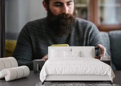 Young millennial man with a long beard sitting on the sofa buys items online with a credit card - Hipster browses the site of his bank to check the balance Wall mural