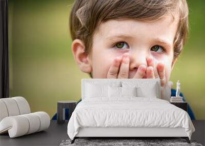 A beautiful little Caucasian boy, looking scared or guilty, covers his face with his hands, hiding, in a park, as if to apologize after having made a little mistake. Concept of emotions in children. Wall mural